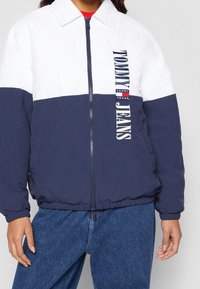 Bicolor jacket with white upper panel and navy lower panel, features a front zipper, logo detailing, and elastic trim on cuffs and hem.