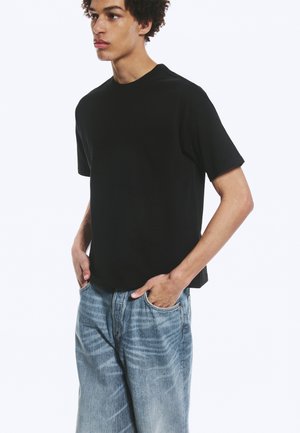 Jonge man met krullend haar, die een effen zwart T-shirt en lichtblauwe spijkerbroek draagt, handen in de zakken, kijkt naar de zijkant.