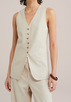 Veste beige sans manches à boutons, associée à un pantalon large assorti, accessoirisée de deux bracelets foncés au poignet droit.