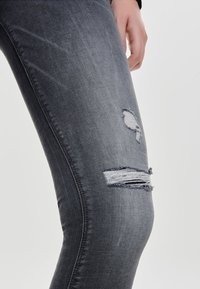 Primer plano de una pierna que lleva pantalones rotos de mezclilla gris oscuro, con agujeros deshilachados por encima y en la rodilla, y una mano descansando al lado del muslo.