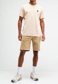 Lichtbeige katoenen T-shirt met een ronde hals en klein zwart logo, gecombineerd met tan katoenen shorts, witte sokken en beige sneakers.