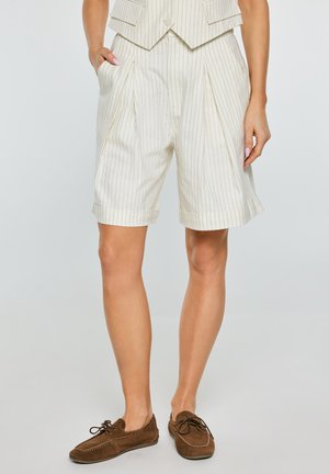 Kobieta ubrana w kremowe, wysokie szorty w prążki z przednimi zakładkami, kieszeniami, podwiniętymi nogawkami i brązowymi mokasynami na sznurowadła.