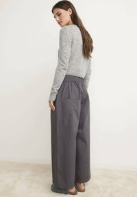 Vrouw met lang bruin haar die een lichtgrijze sweater en losse donkergrijze broek draagt, staand op een beige tapijt tegen een witte muur.