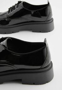 Chaussures en cuir verni noir avec une semelle épaisse, dotées de bouts arrondis, de lacets et d'une finition élégante. Coutures discrètes sur les côtés.
