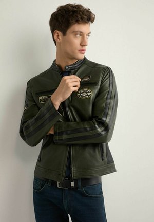 Młody mężczyzna w ciemnozielonej skórzanej kurtce z czarnymi paskami i naszywkami, patrzący w bok z prawą ręką przy kołnierzu.