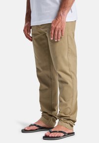 Men's khaki chinos, cotton material, tapered fit, side seam pockets, worn with black flip-flops, relaxed styling, minimal detailing.