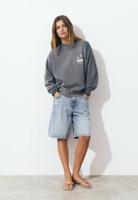 Mujer con cabello largo vistiendo un sudadera gris oversized, pantalones cortos de mezclilla claros hasta las rodillas y chanclas beige, de pie contra una pared blanca.