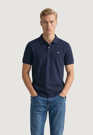 Uomo biondo che indossa una polo blu navy con logo e jeans blu, in piedi con le mani nelle tasche su uno sfondo chiaro e semplice.