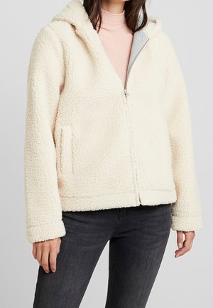 Femme portant une veste crème duveteuse à fermeture éclair avec une capuche, associée à un haut rose clair et un jean gris foncé.