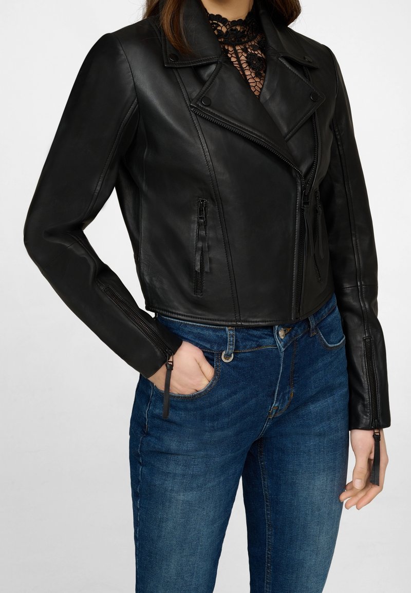 Chaqueta recortada de cuero negro con solapa muescada, frente con cremallera y dos bolsillos con cremallera. Llevada sobre unos jeans de tiro alto azules.