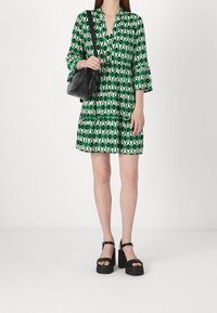 Vihreä kuvioitu mekko, jossa on kolmiomaisia kuvioita, pitkät hihat, V-pääntie ja kerroksellinen yksityiskohta. Yhdistetty mustan laukun ja paksupohjaisten mustien kenkien kanssa.