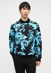 Άνδρας που φορά μαύρο πουκάμισο με μακριά μανίκια, με φωτεινά μπλε κυματιστά μοτίβα νερού και μαύρο γιακά, στέκεται μπροστά σε απλό φωτεινό φόντο.