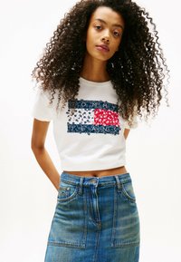 T-shirt blanc cropped avec un motif floral bleu et rouge, portant le texte "TOMMY JEANS". Accompagné d'une jupe en denim évasée, de lavage moyen.
