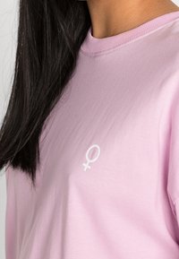 T-shirt a maniche lunghe di colore rosa chiaro realizzato in tessuto morbido, con un simbolo di genere femminile bianco sul petto e design a girocollo.