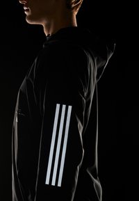 Svart huva jacka med texturerad tyg. Har reflekterande tre-stripes design på ärmen. Enkel, modern snitt mot en mörk bakgrund.