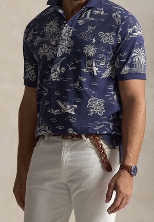 Homme portant un polo bleu marine avec des imprimés tropicaux et nautiques blancs, un pantalon blanc avec une ceinture en cuir tressé marron et une montre en cuir marron.