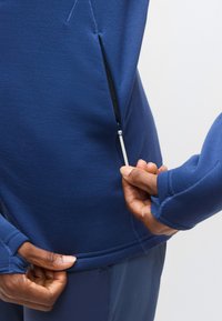 Αθλητικό μπλούζα σε ναυτικό μπλε με τσέπη με φερμουάρ, απαλό ύφασμα, εφαρμοστά μανσέτες και τρύπες για τον αντίχειρα για αυξημένη άνεση και λειτουργικότητα.