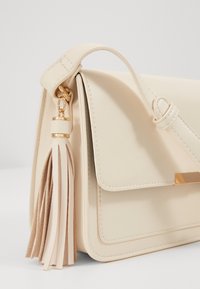 Krémsvětlá kožená kabelka s klopou na zapínání, vybavená zlatými kovovými prvky a dlouhým střapcem na boku. Hladký povrch a minimalistický design.