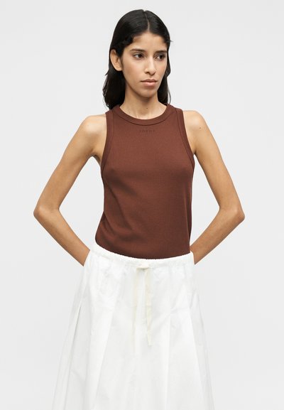 Καφέ τοπ χωρίς μανίκια με ριγέ υφή και στρογγυλή λαιμόκοψη, σε συνδυασμό με λευκή φούστα με κορδόνι στη μέση. Απλός και εφαρμοστός σχεδιασμός.