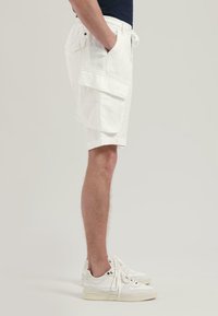 Weiße Cargoshorts aus strukturiertem Stoff, mit einem Kordelzugbund, seitlichen Taschen und gestickten Details, kombiniert mit weißen Sneakers.