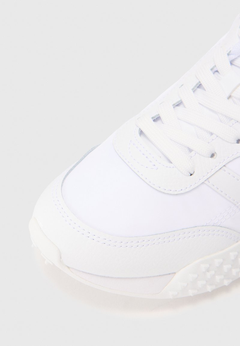 Zbliżenie na czubek białego trampka z teksturowaną podeszwą i sznurówkami na jasnoszarym tle.