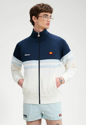 Młody mężczyzna w granatowo-białej kurtce zapinanej na zamek i jasnoniebieskich szortach, stojący z rękami w kieszeniach, patrzący w bok.