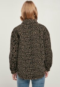 Kobieta z blond włosami, ubrana w brązowo-czarną futrzaną kurtkę w panterkę z długim rękawem, zestawioną z niebieskimi dżinsami, pokazana od tyłu.