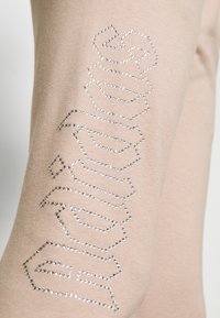 Beige legingi ar izteiktu kristalizētu logotipa detaļu sānos, ar gludu tekstūru un elastīgu audumu.