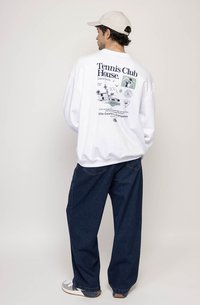 Sweatshirt branco com desenhos gráficos nas costas, combinado com jeans igualmente soltos de cor azul escura e ténis de tom claro.