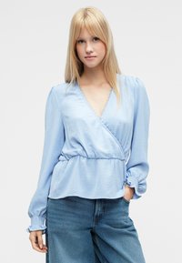 Lyseblå slå-om bluse med lange ærmer, flæsekantede manchetter og struktureret materiale. Har en V-udskæring med flæsekant og en elastisk talje.