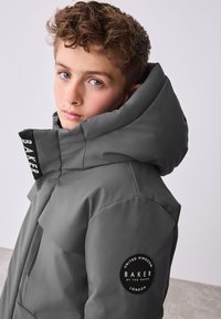 Hall beežist topeltpolsterdatud jope, millel on suur pehme kapuuts. Vasakul varrukal on must logomärk ja krael must silt. Sile tekstuur.