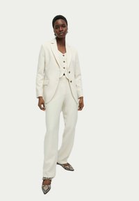 Completo pantalone bianco su misura caratterizzato da un blazer aderente con rever e pantaloni abbinati. La parte superiore ha tre bottoni marroni e una texture liscia.