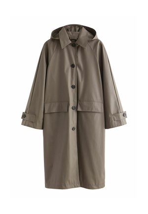 Personne portant un long trench-coat gris, un haut noir, un pantalon blanc et portant un sac à bandoulière noir texturé dans un espace intérieur lumineux.