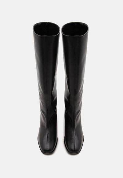 Paire de bottes hautes en cuir noir avec des bouts carrés, debout côte à côte sur un fond clair uni.