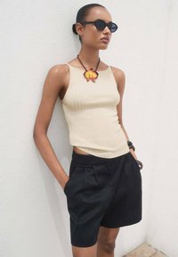 Top morbido a coste di colore crema, pantaloni corti neri sartoriali con pieghe e una collana con pendente a forma di fiore multicolore. Modella che indossa occhiali da sole rotondi neri.