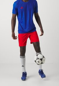 Blå atletisk T-shirt med röda shorts, vita knä-långa strumpor med svarta ränder, blå fotbollsskor och en svartvit fotboll.