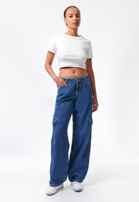 Camiseta blanca cropped y pantalones cargo de pernera ancha azul con dos bolsillos laterales. Los pantalones tienen un cierre de botón y están hechos de tela de denim.