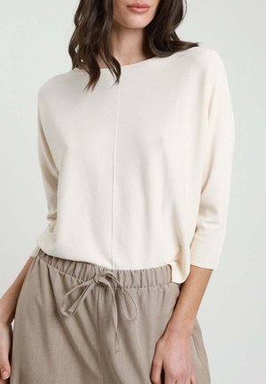 Donna che indossa un morbido maglione crema a maniche tre quarti infilato in pantaloni beige con coulisse, su sfondo semplice.