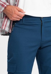 Pantaloni cargo blu realizzati in tessuto elasticizzato, con tasche con zip, una cintura ampia e una tasca con flap sulla coscia. Dettaglio testurizzato sul tessuto.