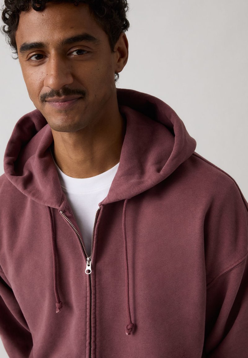 Man met kort krullend haar en snor, gekleed in een bordeauxrode hoodie met rits over een wit shirt tegen een effen achtergrond.