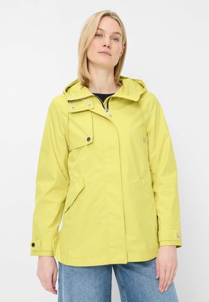 Frau trägt eine hellgrüne Kapuzenjacke mit Reißverschluss- und Knopfdetails, kombiniert mit blauen Jeans, steht vor weißem Hintergrund.