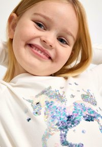 Hvid langærmet top med et farverigt paliet-unicorn design med blå, grønne og sølv detaljer, som har en glat tekstur.