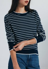 Marinblå randig tröja med vita linjer, ribbad halsringning och muddar, med guldknappsdetaljer på ärmarna. Mjuk stickad tyg.