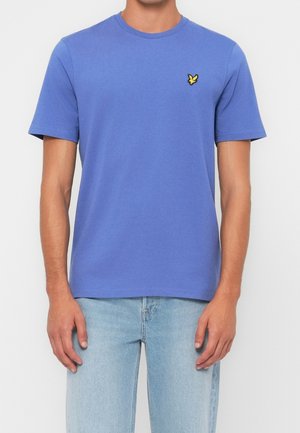 Mand iført en ensfarvet blå kortærmet t-shirt med et lille gult fuglelogo på venstre bryst og lyseblå jeans.