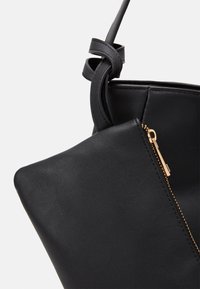 Gros plan d’un sac à main en cuir noir avec un détail de sangle nouée et une fermeture éclair dorée sur une petite pochette attachée.