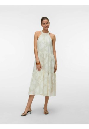 Moteris vilki lengvą gėlėtą midi suknelę be rankovių ir aukštakulnius sandalus, stovi prieš baltą foną ir šypsosi.