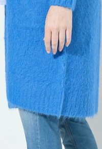 Flauschige blaue Strickjacke mit weicher Textur, die einen geraden Schnitt aufweist. Eine Hand ruht sanft auf dem Stoff und hebt die Farbe und das Material hervor.