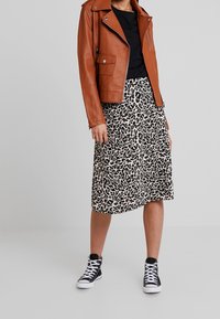 Giacca da moto in pelle marrone, maglietta nera, gonna midi a stampa leopardata e sneakers alte nere. La texture varia tra i diversi capi.