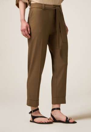 Pantalons marron ajustés avec taille ceinturée, jambes droites et ourlets retroussés, associés à des sandales plates noires avec brides de cheville.