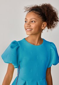 Das blaue Kleid hat Puffärmel und ist mit wiederholten 'B'- und Schleifenmustern strukturiert. Kontraststreifen an den Seiten fügen dem Design detaillierte Elemente hinzu.